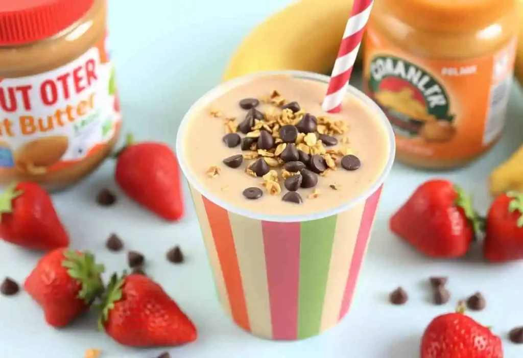 A colorful cup of creamy peanut butter fruit smoothie from kid-friendly smoothie recipes, topped with granola and mini chocolate chips, with fresh strawberries, bananas, and a jar of peanut butter nearby, on a bright and cheerful background.