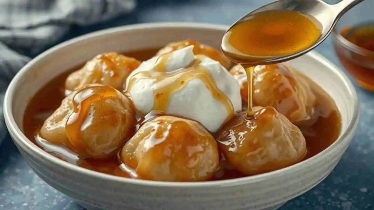 A plate of golden syrup dumplings lightly drizzled with caramel sauce, emphasizing the soft, fluffy texture. A dollop of cream tops the dumplings for added richness. A silver spoon rests nearby, completing the cozy dessert presentation.