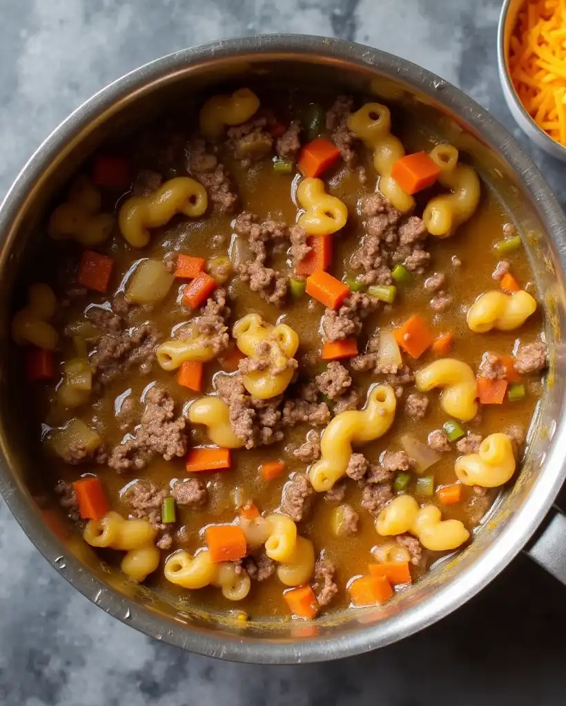 A pot on the countertop containing ground beef, sautéed onions, carrots, celery, and simmering elbow macaroni in a creamy broth, alongside a bowl of shredded cheddar 