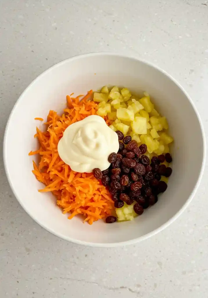 10-Minute Summer Carrot Raisin Salad 3 Grated carrots, soaked raisins, and pineapple tidbits being added to a creamy dressing in a white bowl for a summer carrot raisin salad.