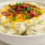 A homemade mashed potato bowl topped with cheese, crispy turkey bacon, and green onions, served in a white bowl on a kitchen countertop.