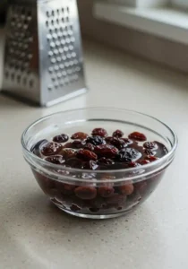 Freshly grated orange carrots next to a bowl of dark raisins soaking in water, key ingredients for a summer carrot raisin salad.