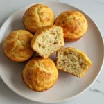A white plate holds three golden-brown cottage cheese banana muffins, with one sliced in half to show its soft, moist crumb.
