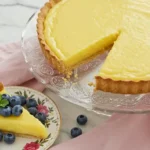 A perfect slice of homemade lemon tart on a white plate, showing the thick yellow curd filling and buttery crust, with a dusting of powdered sugar.