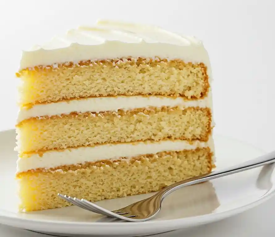 Simple Vanilla Sponge Cake 3 A slice of vanilla sponge cake on a white plate with a fork. The slice shows the golden cake and layers of smooth.
